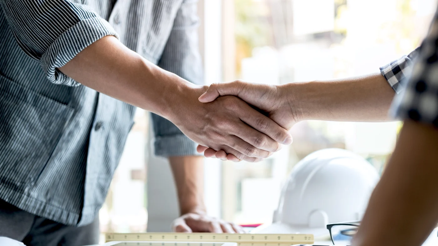 Standard Setting Roof Replacement Austin Area Willi Roofing Services Close view of two men shaking hands over a contractor’s table with a hard hat, ruler, and paperwork sitting on it