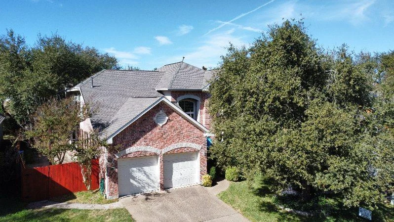 An aerial view of a new asphalt shingle roof on a two-story brown house with white garage doors.