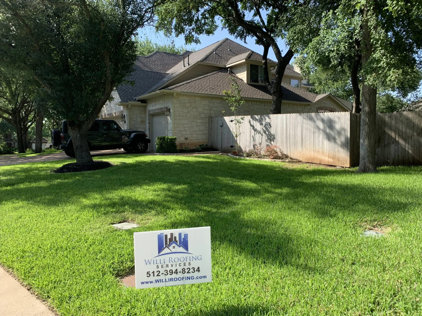 Two-story stone and stucco home with a brown asphalt shingle roof surrounded by lush green grass and mature trees.