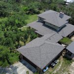 Aerial view of a large residential home with an asphalt shingle roof, surrounded by dense green trees, multiple roof sections, and an attached driveway.