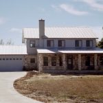 Front exterior view of a two-story stone home with a newly installed standing seam metal roof in Kyle, TX, featuring a covered porch