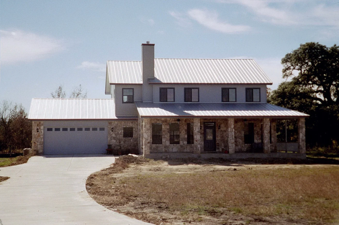 Seamless Metal Roof Replacement Kyle Tx Willi Roofing Front exterior view of a two-story stone home with a newly installed standing seam metal roof in Kyle, TX, featuring a covered porch