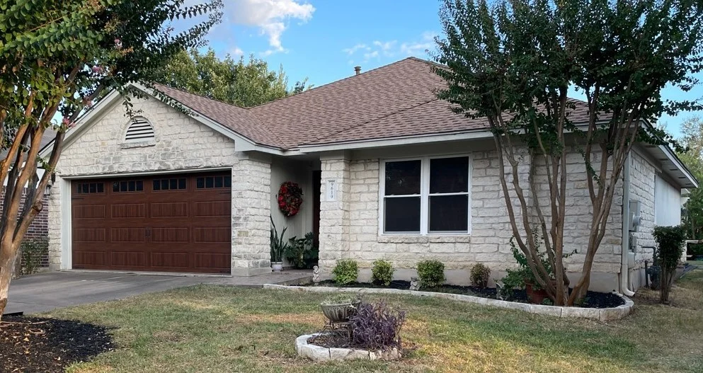 Single-story stone home featuring a new light-brown asphalt shingle roof, wooden garage door, trimmed landscaping, and a small front porch.