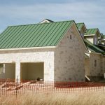A newly constructed stone house with a green standing seam metal roof, shown behind orange safety fencing at an active construction site in Dripping Springs, TX.