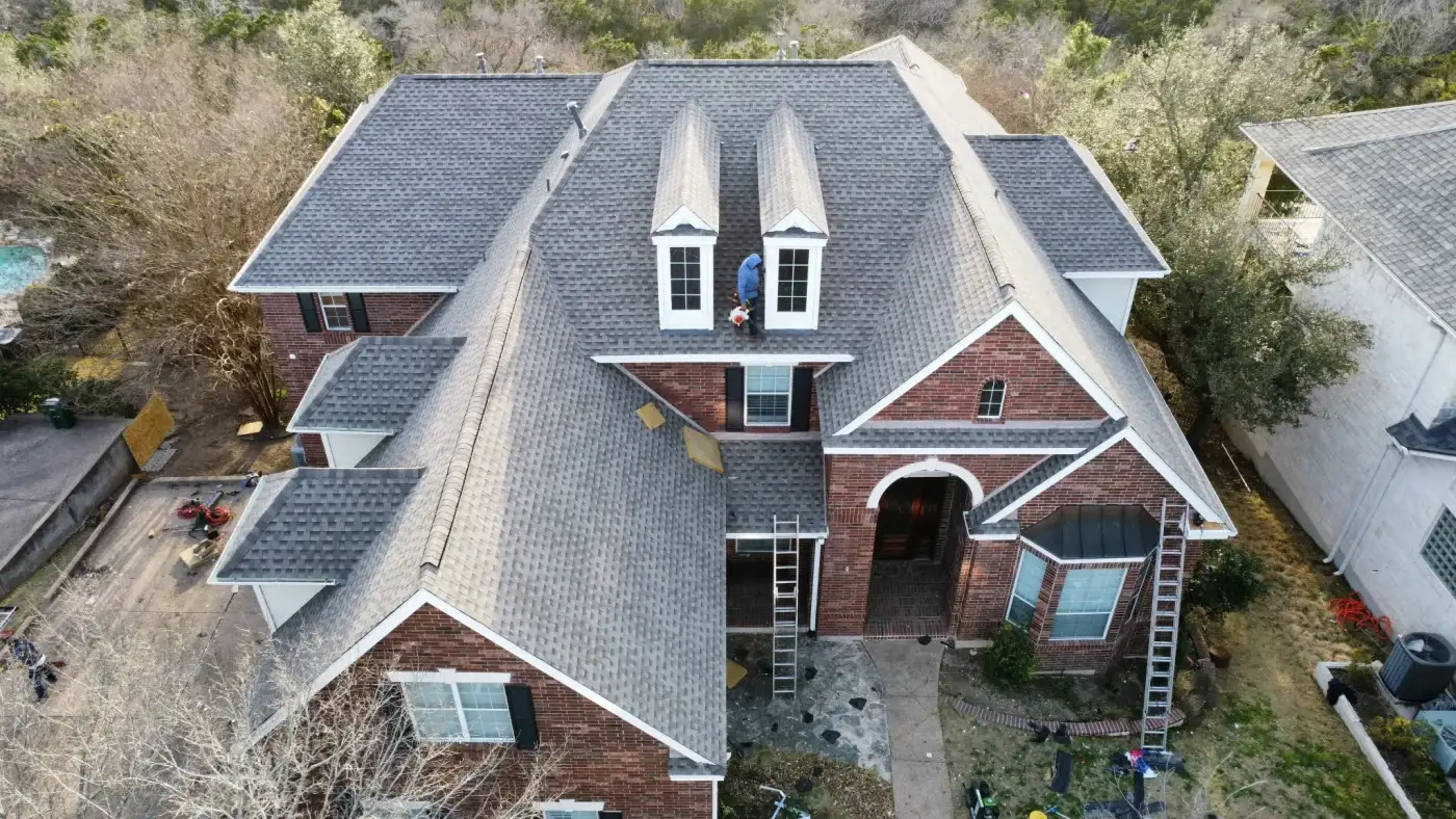 A single-story home with brick siding, a gray shingle roof, and trees in the front yard.